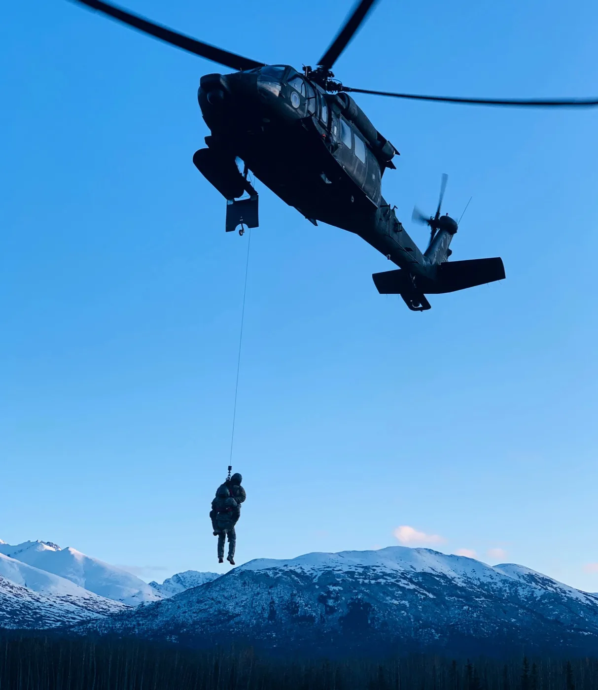 A Black Hawk helicopter hoists a soldier over snow-capped Alaska mountains at dusk