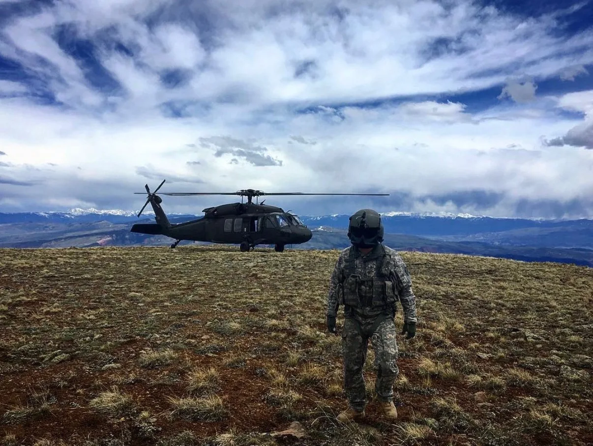 A hoist operator standing before a landed Black Hawk on an Alaska mountain ridge